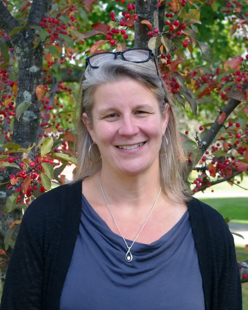 chloe wears a blue shirt and black cardigan stanging against a red flowering tree