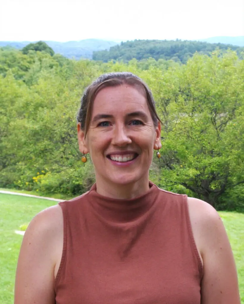 Emily wears a terra cotta colored sleeveless shirt and stands against a green field and mountains in the background