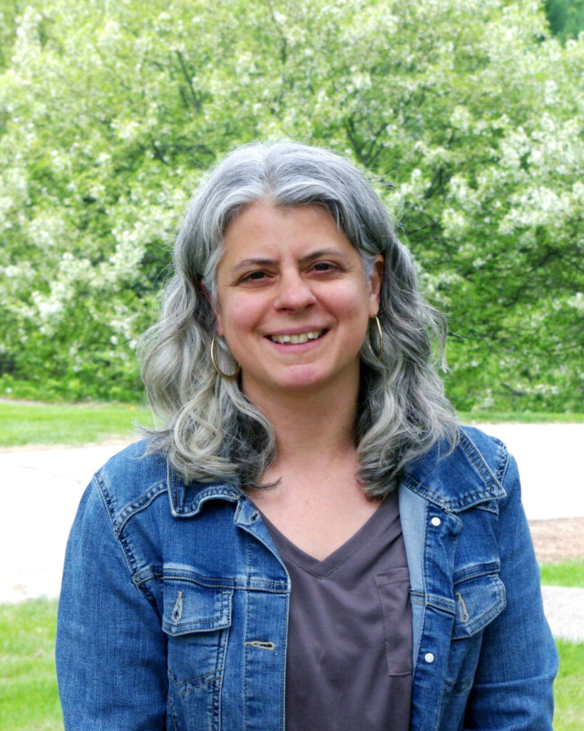 Sarah wears a grey shirt under an open denim shirt against a green background