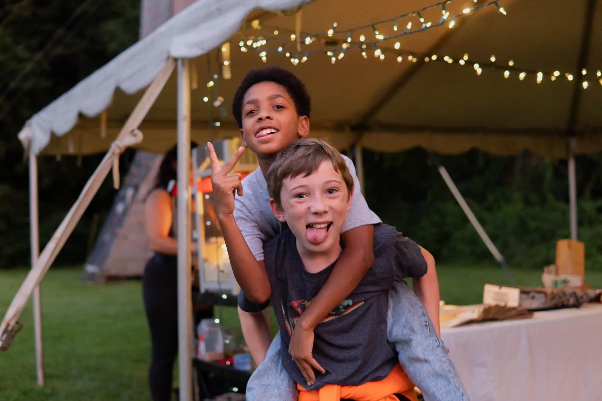 One kid is carrying another on their back. Both are making silly faces at the camera and standing against a backdrop of twinkle lights