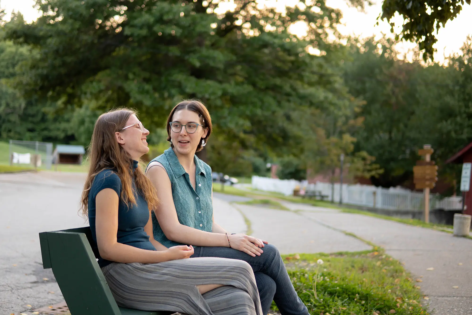 Two friends sit on a bench and laugh together as the sun sets behind some trees in the distance