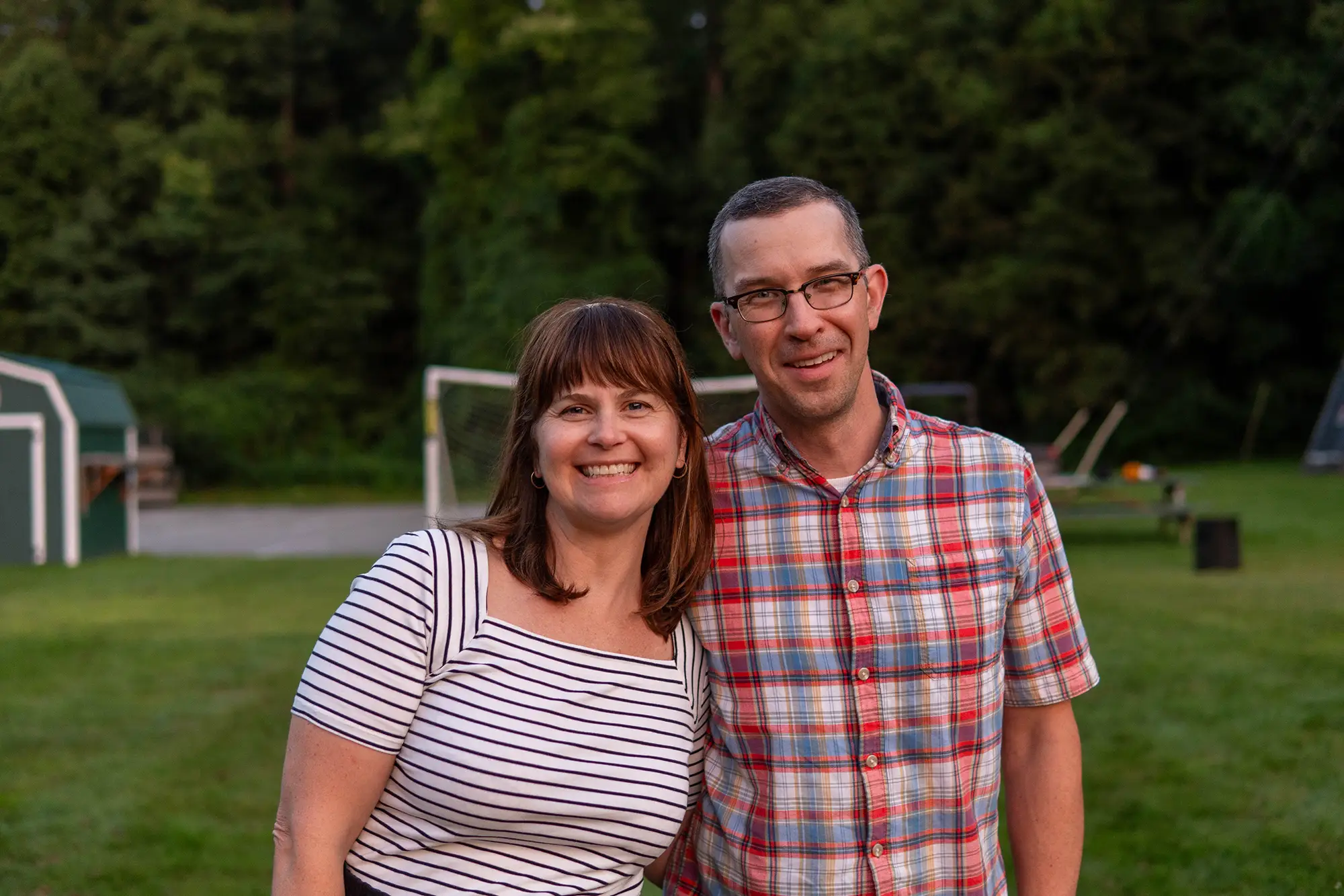 A couple standing on a soccer field smiles at the camera wearing stripes and plaid